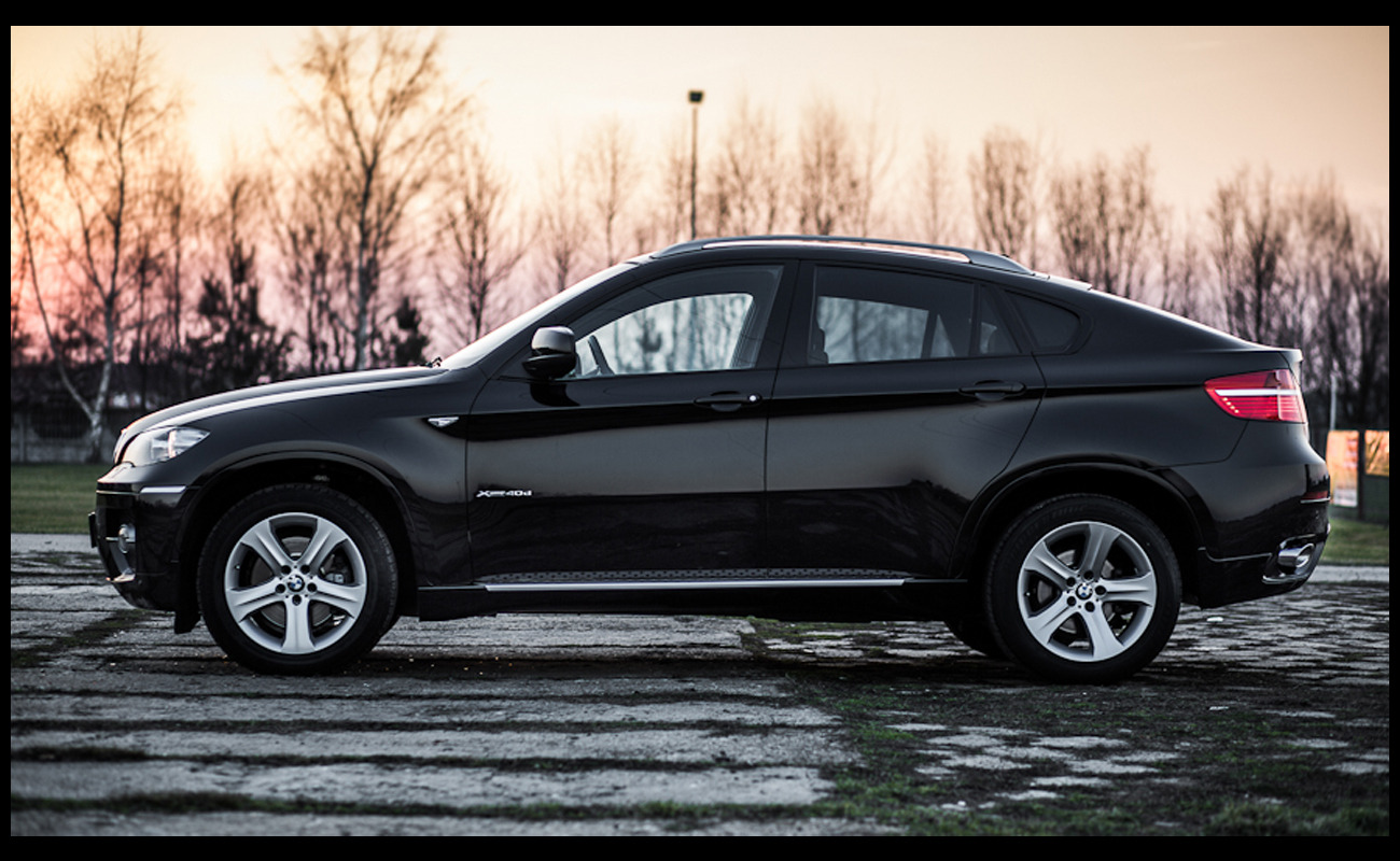 Czarny samochód BMW X6 xDrive40d, widok z boku na tle drzew o zachodzie słońca, na betonowej nawierzchni z elementami zieleni.