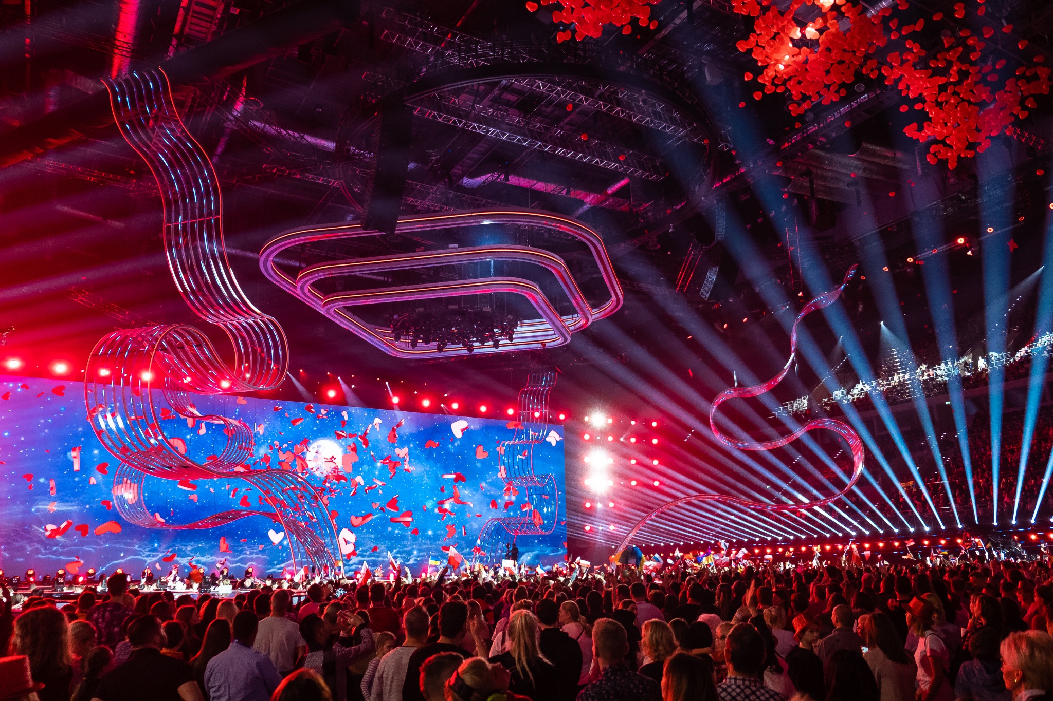 Widok na scenę podczas dużego wydarzenia z tłumem ludzi, efektowną scenografią z metalowymi konstrukcjami, oświetleniem punktowym i ekranem LED wyświetlającym animacje serc.
