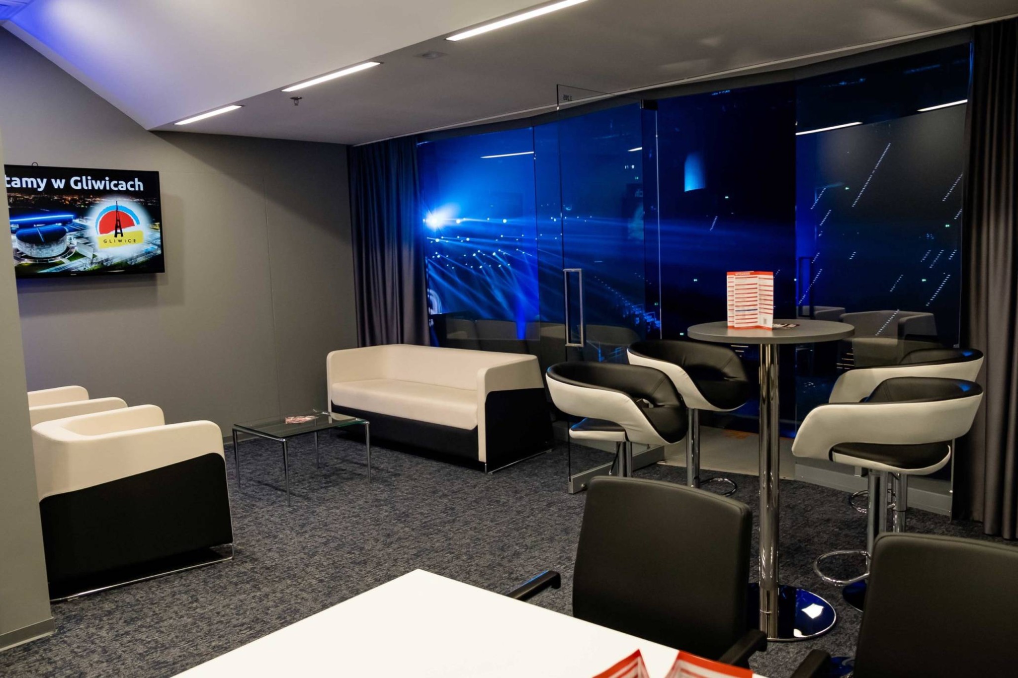 Nowoczesne wnętrze sali konferencyjnej z białymi sofami, krzesłami barowymi i szklaną ścianą z widokiem na oświetloną scenę widowiskową w tle.