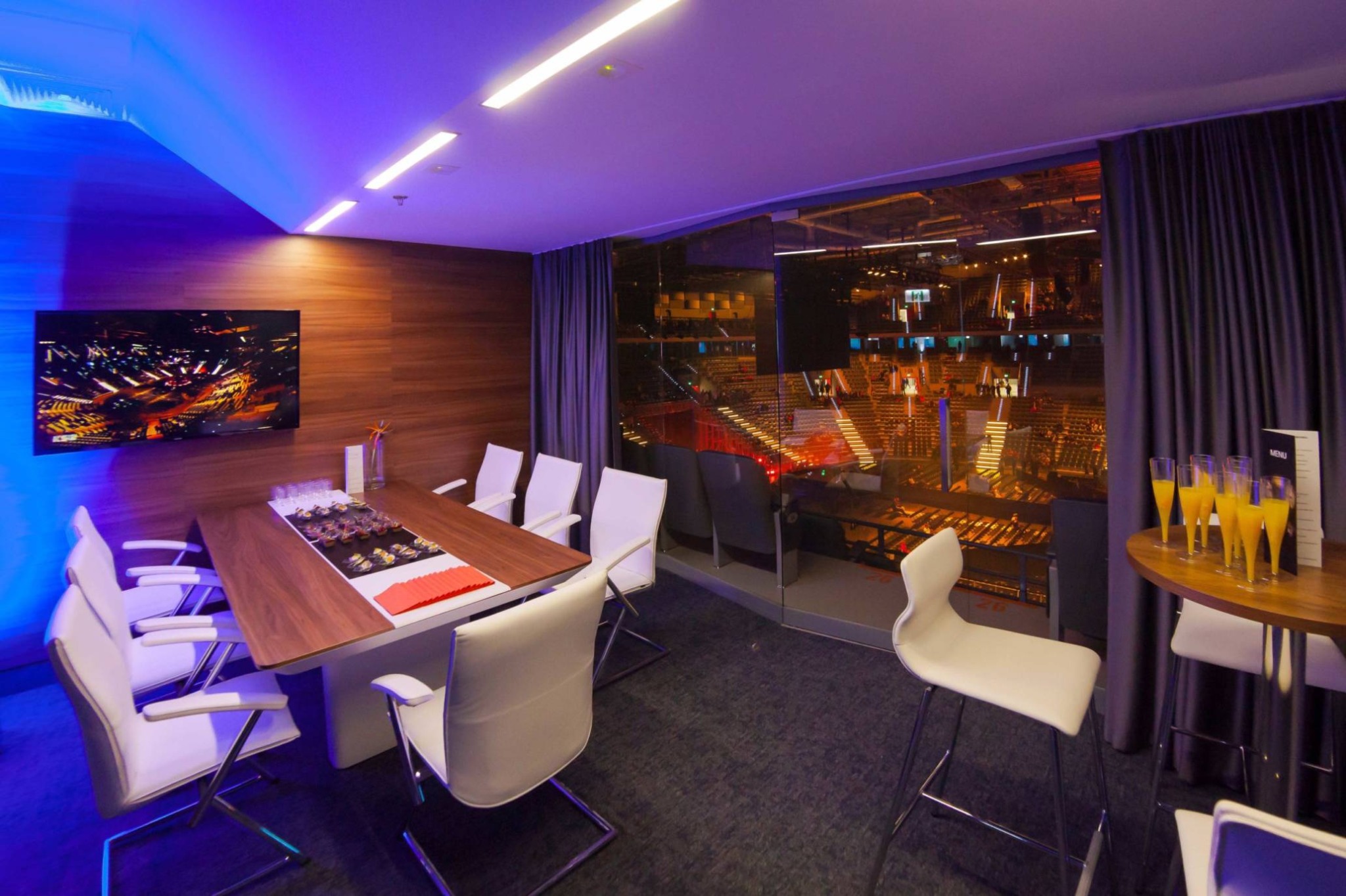 Loża VIP z widokiem na arenę, stół z przekąskami i napojami, nowoczesne oświetlenie, krzesła konferencyjne.