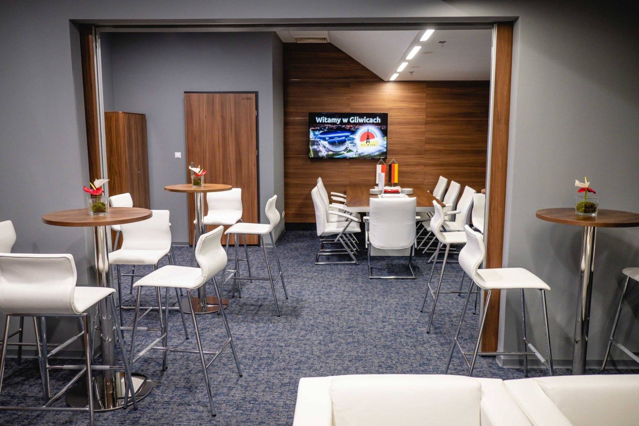Sala konferencyjna z wysokimi stołami barowymi, białymi krzesłami, długim stołem konferencyjnym i telewizorem wyświetlającym panoramę stadionu w Gliwicach.