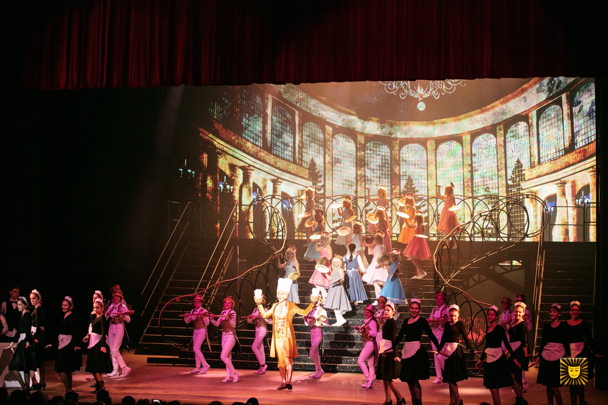 Scena teatralna z grupą aktorów w kostiumach, w tle wyświetlana cyfrowo scenografia pałacowa z kolumnami i schodami.