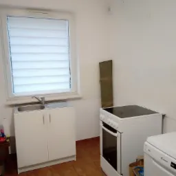 Wnętrze małej kuchni z białymi meblami: szafka pod zlewem, kuchenka elektryczna, pralka. Na podłodze brązowe płytki. Okno z żaluzjami poziomymi.