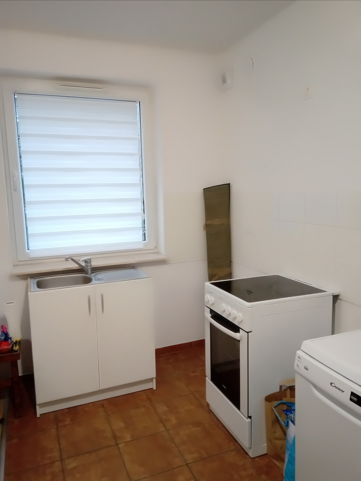 Wnętrze małej kuchni z białymi meblami: szafka pod zlewem, kuchenka elektryczna, pralka. Na podłodze brązowe płytki. Okno z żaluzjami poziomymi.