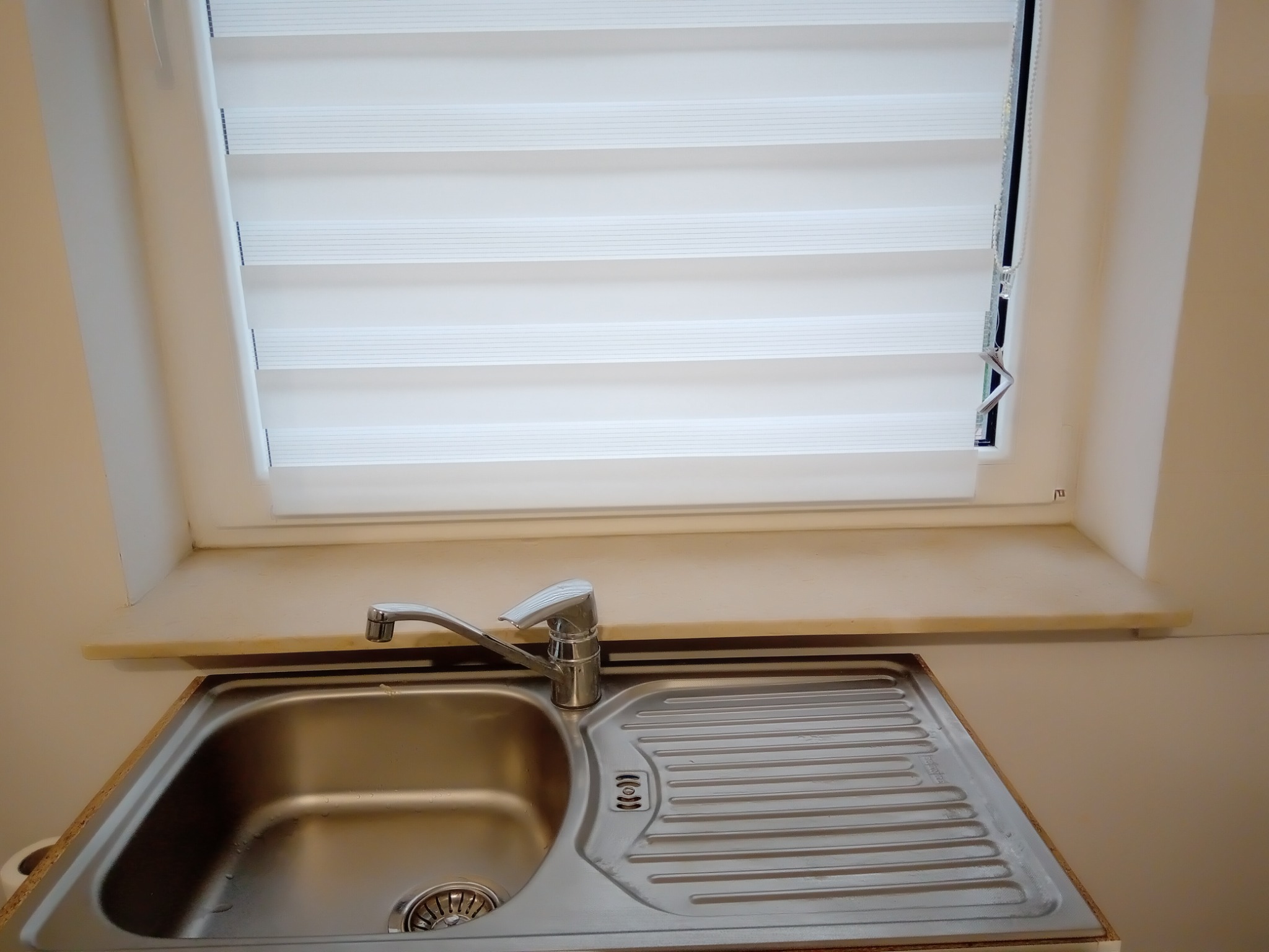 Stalowy zlew jednokomorowy z ociekaczem i baterią, zamontowany pod oknem z roletami dzień-noc.