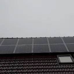 Panele słoneczne na brązowym dachu z dachówkami, zamontowane nad oknami dachowymi, widok z dołu w pochmurny dzień.