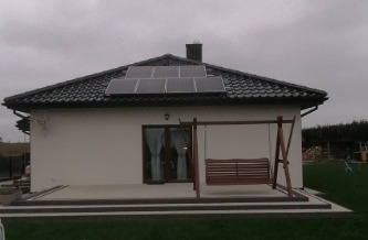 Dom jednorodzinny z zamontowanymi panelami fotowoltaicznymi na dachu pokrytym szarą dachówką, widoczne z boku, z huśtawką ogrodową na pierwszym planie.