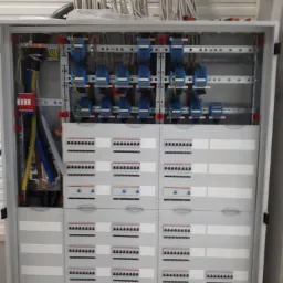 Otwarta skrzynka elektryczna z widocznymi bezpiecznikami, przewodami i listwami montażowymi wewnątrz, z licznymi kablami ułożonymi nad nią.