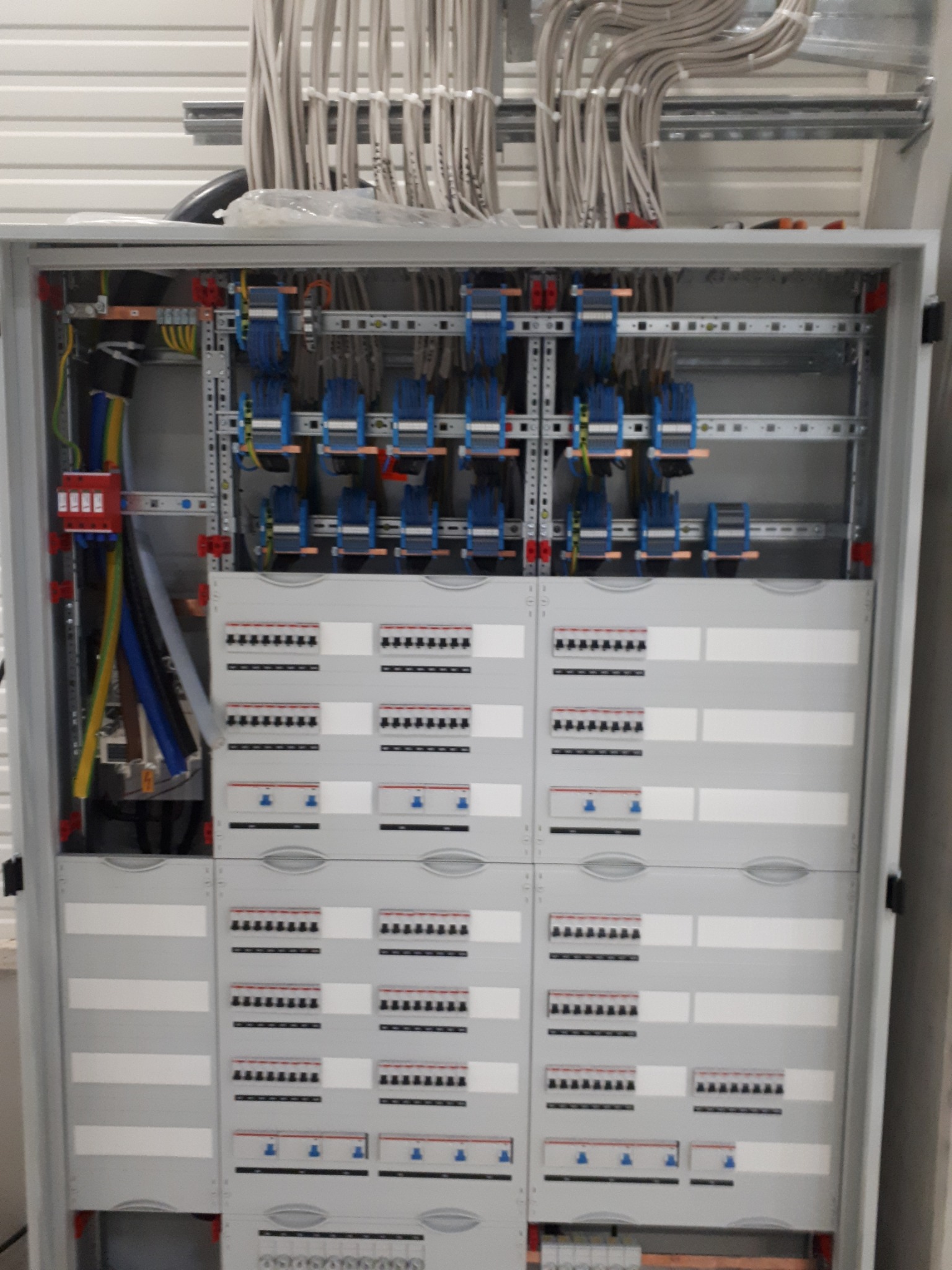 Otwarta skrzynka elektryczna z widocznymi bezpiecznikami, przewodami i listwami montażowymi wewnątrz, z licznymi kablami ułożonymi nad nią.