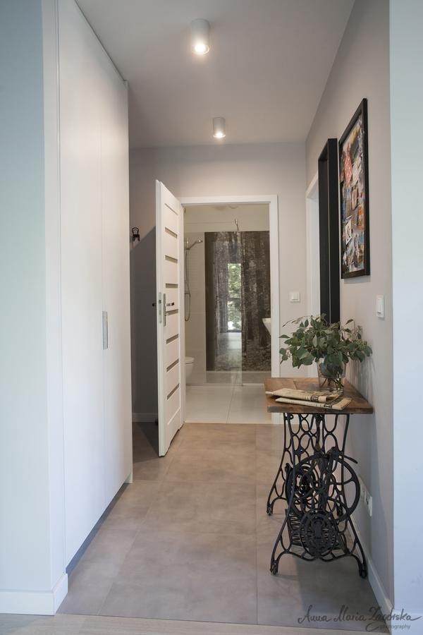 Nowoczesny przedpokój z szarą podłogą, białymi szafami i otwartymi drzwiami do łazienki z fototapetą leśną. Na pierwszym planie stolik na bazie starej maszyny do szycia z bukietem zielonych liści.