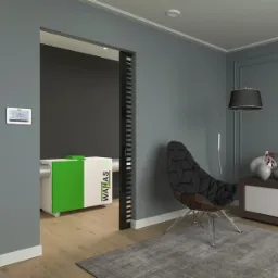 Biało-zielony rekuperator WANAS umieszczony w pomieszczeniu mieszkalnym, widoczny przez otwarte drzwi, obok nowoczesny fotel i lampa, na ścianie panel sterowania.