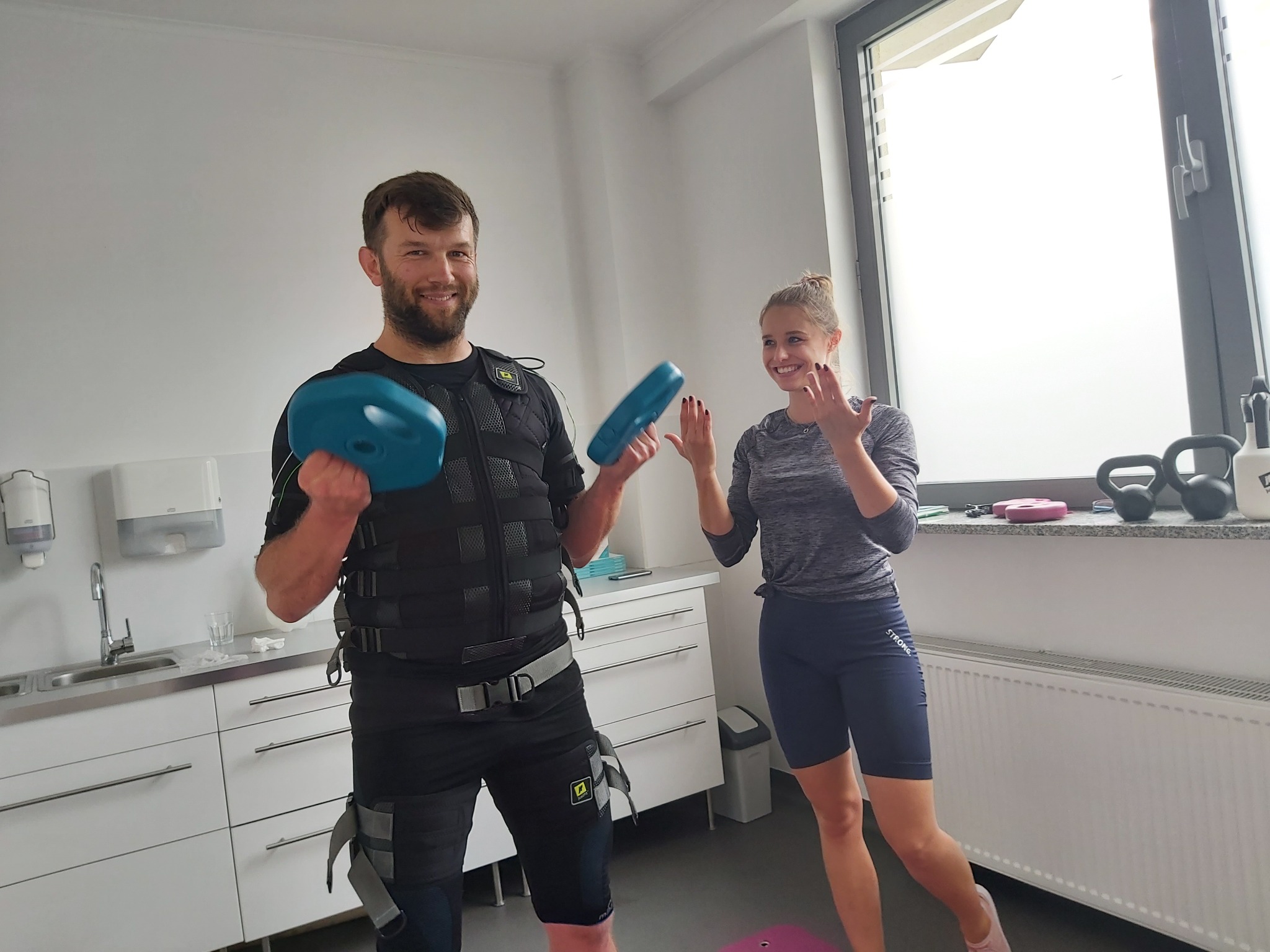 Mężczyzna z uśmiechem na twarzy, ubrany w kamizelkę do elektrostymulacji mięśni, trzymający w dłoniach elektrody, obok niego uśmiechnięta kobieta w sportowym stroju, w tle białe meble i okno.