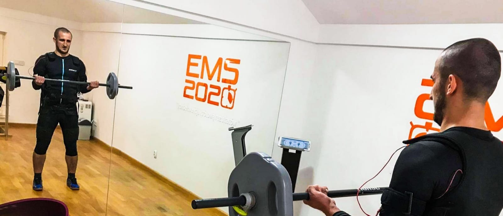 Mężczyzna w kombinezonie EMS ćwiczy biceps z obciążeniem na gryfie w siłowni z lustrami i logo EMS 2020 na ścianie.