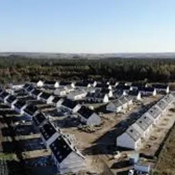 Osiedle domów jednorodzinnych z dachami pokrytymi szarą dachówką, widok z lotu ptaka, w tle las i pole.