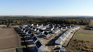 Osiedle domów jednorodzinnych z dachami pokrytymi szarą dachówką, widok z lotu ptaka, w tle las i pole.