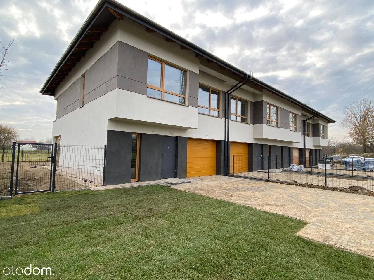 Nowoczesne segmenty mieszkalne z elewacją w odcieniach bieli i szarości, pomarańczowe bramy garażowe, świeżo założony trawnik i ogrodzenie z siatki.