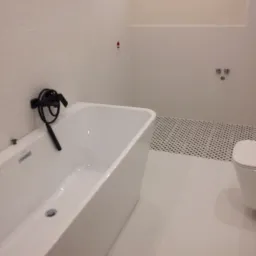 Biała, wolnostojąca wanna z czarną armaturą na tle białych płytek ściennych z delikatną fakturą i podłogi z białych płytek oraz fragmentu podłogi z mozaiką w czarno-białą szachownicę w nowoczesnej...