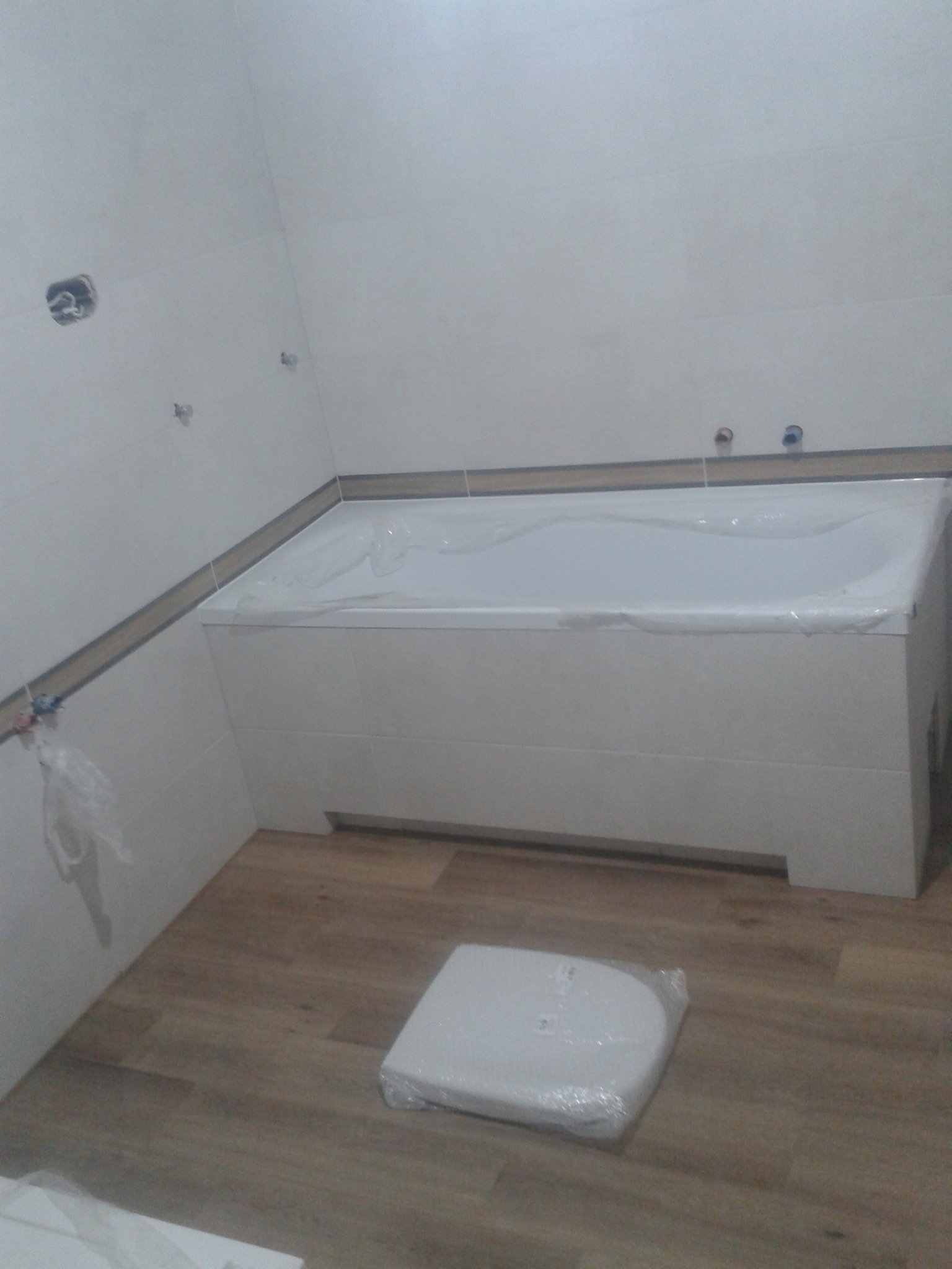 Nowa, biała wanna obudowana płytkami, zabezpieczona folią ochronną w łazience z drewnopodobną podłogą i fragmentem instalacji elektrycznej na ścianie.