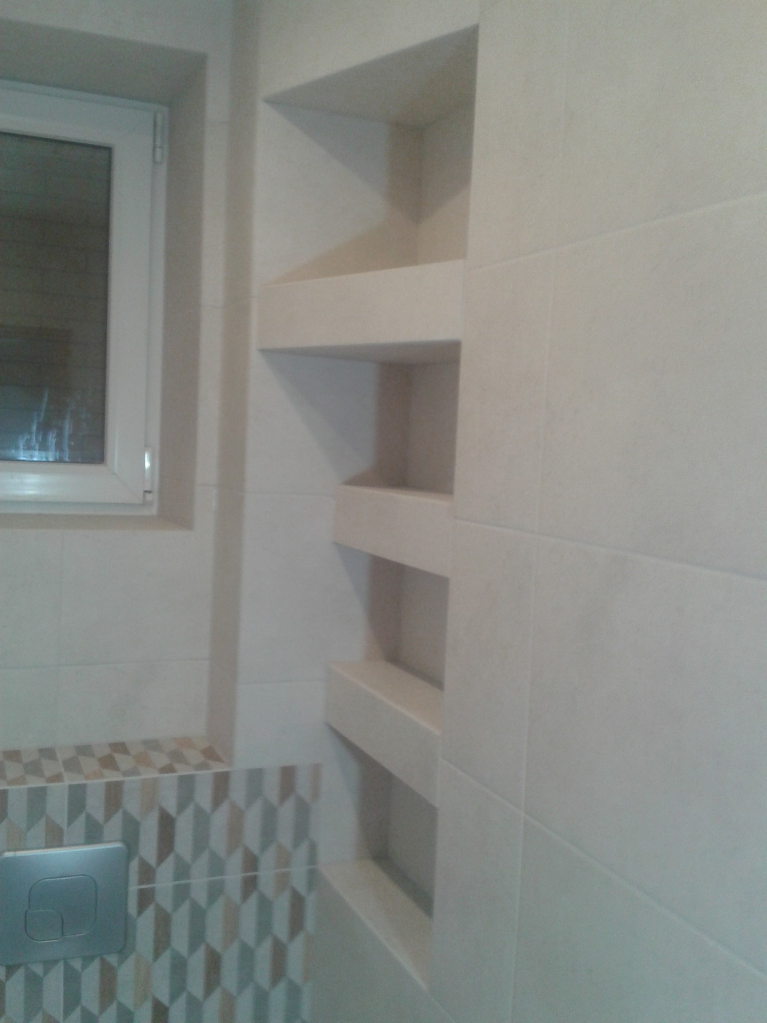 Wykończona łazienka z wnęką półkową wyłożoną płytkami, obok okno i ozdobny pas płytek z przyciskiem spłuczki.