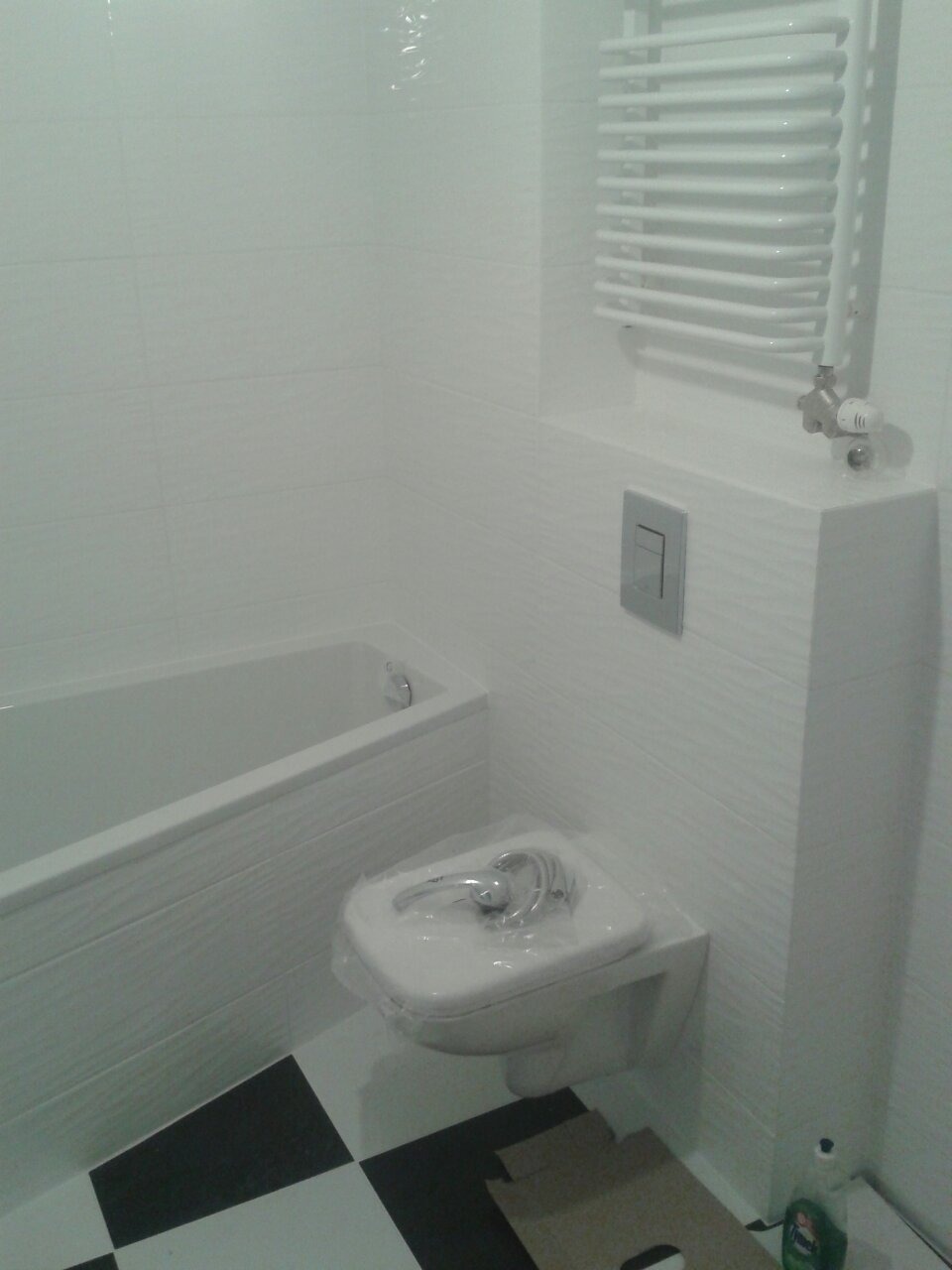 Nowa, biała toaleta w folii ochronnej, zamontowana w łazience w Łodzi z białą wanną i grzejnikiem drabinkowym na ścianie z białymi płytkami o fakturze drewna, czarno-biała podłoga w szachownicę.