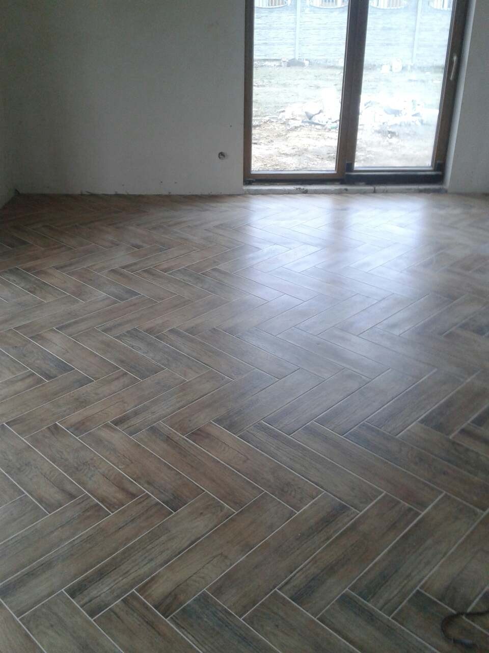 Ułożona podłoga z brązowych płytek imitujących drewno w jodełkę, widok na okno balkonowe z białym płotem w tle.