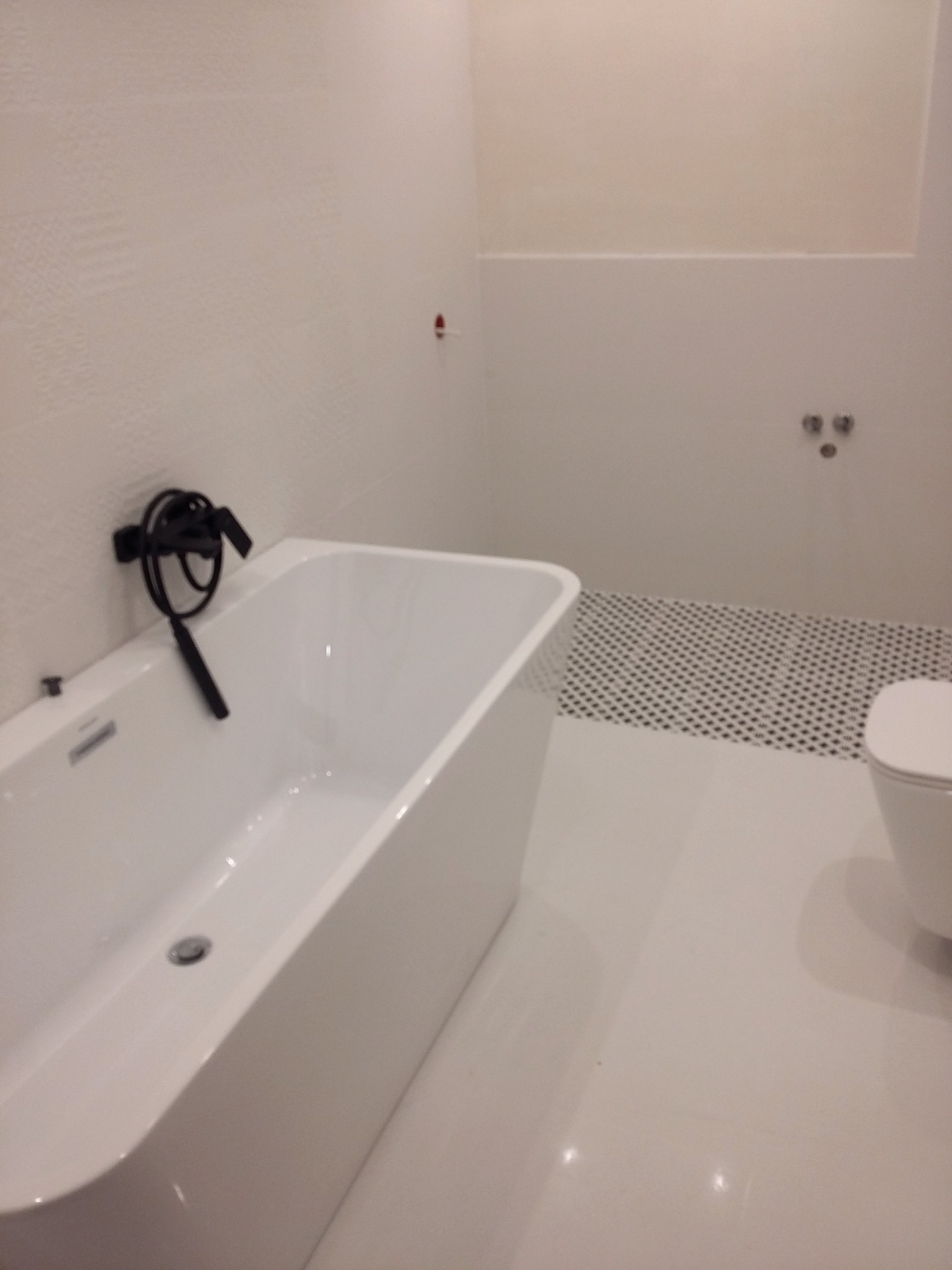 Biała, wolnostojąca wanna z czarną armaturą na tle białych płytek ściennych z delikatną fakturą i podłogi z białych płytek oraz fragmentu podłogi z mozaiką w czarno-białą szachownicę w nowoczesnej...