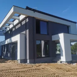 Nowoczesny dom dwukondygnacyjny z szarą elewacją i białymi elementami architektonicznymi, widoczne niedokończone prace wykończeniowe wokół budynku, teren nierówny z piaskiem i śladami opon.