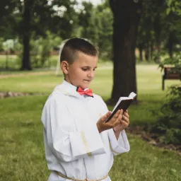 Chłopiec w albie komunijnej z czerwoną muchą czyta modlitewnik na tle zielonego trawnika i drzew
