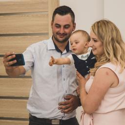 Fotografia i filmowanie Adam Sielski - Rodzina robiąca selfie telefonem komórkowym w jasnym wnętrzu: ojciec trzyma telefon, matka trzyma małe dziecko z wyciągniętą rączką, wszyscy uśmiechnięci.