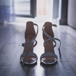 Fotografia i filmowanie Adam Sielski - Eleganckie, srebrne szpilki z brokatowym paskiem, ustawione na ciemnej, drewnianej podłodze w kierunku otwartych drzwi, ukazujące wnętrze mieszkania.