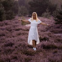Fotografia i filmowanie Adam Sielski - Uśmiechnięta blondynka w białej sukience i sportowych butach spaceruje po polu wrzosów w otoczeniu drzew, z rozłożonymi ramionami, w delikatnym, naturalnym świetle.