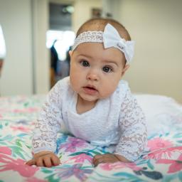 Fotografia i filmowanie Adam Sielski - Urocze niemowlę w białej koronkowej sukience i opasce z kokardą leży na kwiecistej pościeli, patrząc prosto w obiektyw z wyrazem zaciekawienia.