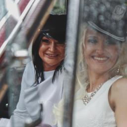 Fotografia i filmowanie Adam Sielski - Dwie uśmiechnięte kobiety w stylizowanych strojach i czapkach siedzą w samochodzie retro, widoczne przez zaparowaną szybę z naklejką 'Wunder-Baum'.