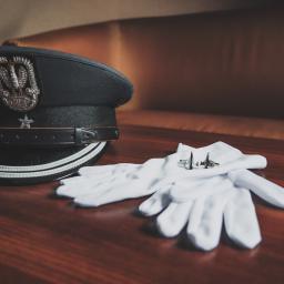 Fotografia i filmowanie Adam Sielski - Czarny kapelusz galowy z godłem, białe rękawiczki i spinki do mankietów w kształcie samolotów na ciemnobrązowym drewnianym stole.