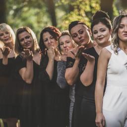 Fotografia i filmowanie Adam Sielski - Panna młoda w białym kombinezonie z szarfą 'Bride to be' i koroną na głowie, pozuje z druhnami w czarnych sukienkach, wysyłając buziaki w plenerze.