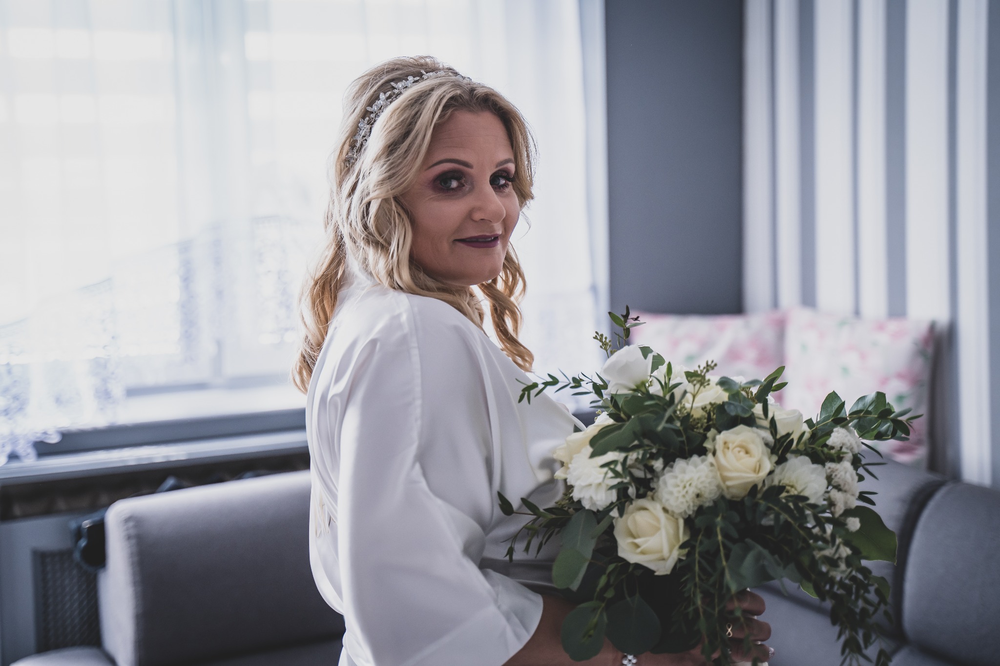 Panna młoda w białym szlafroku, z blond włosami upiętymi ozdobną opaską, trzyma bukiet białych kwiatów na tle okna i szarej kanapy.