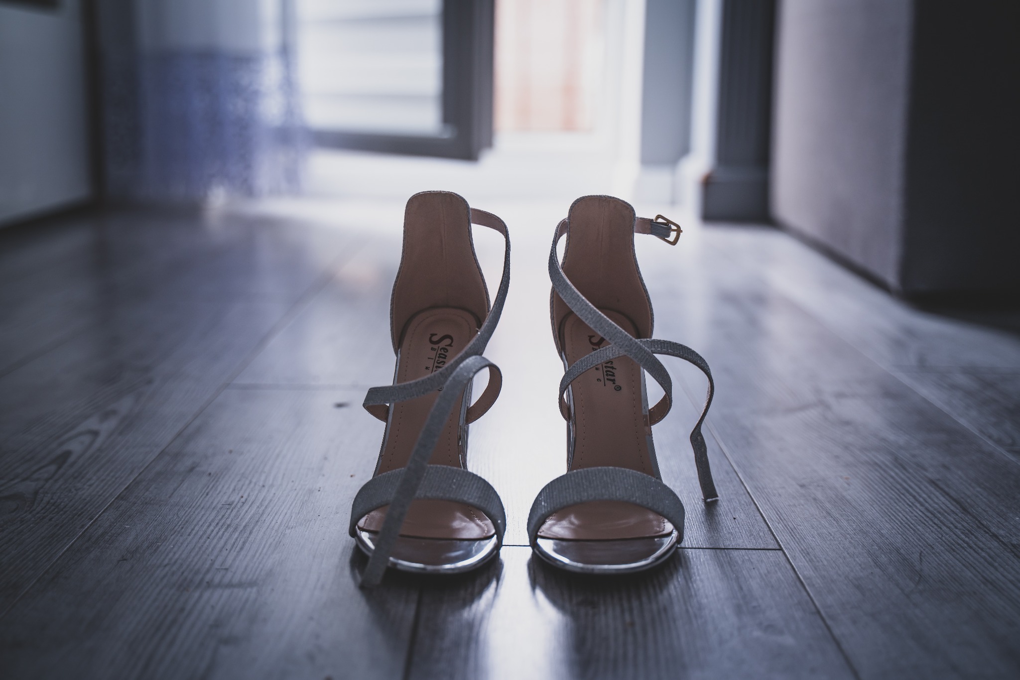 Eleganckie, srebrne szpilki z brokatowym paskiem, ustawione na ciemnej, drewnianej podłodze w kierunku otwartych drzwi, ukazujące wnętrze mieszkania.