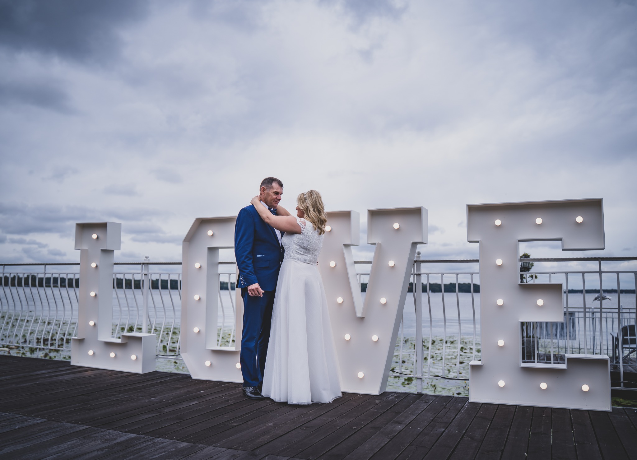 Para młoda w objęciach na drewnianym pomoście, przed dużym napisem LOVE z żarówkami, jezioro w tle, pochmurne niebo.