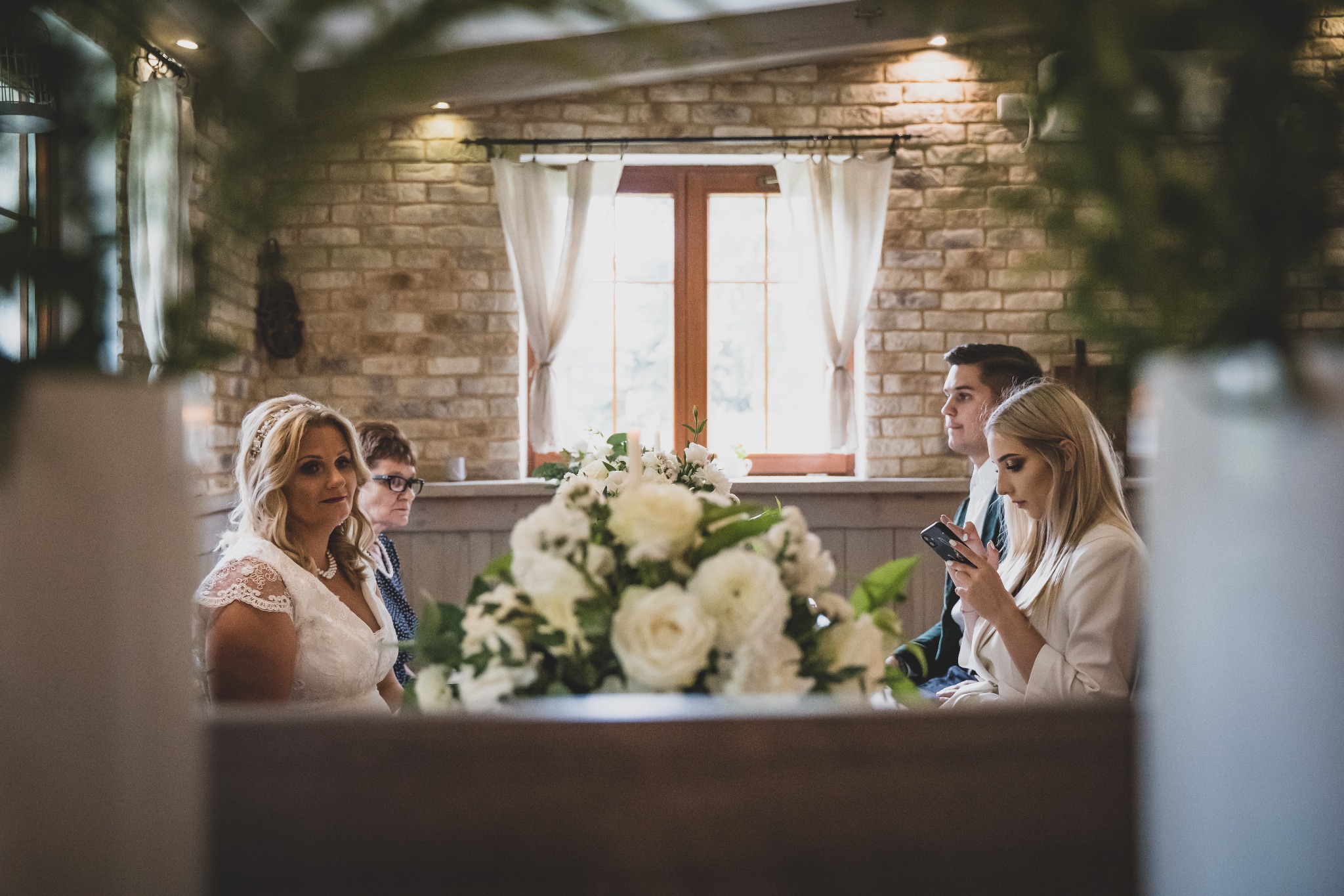 Panna młoda w koronkowej sukni siedzi podczas ceremonii, w tle ceglana ściana i okno z białymi zasłonami, na pierwszym planie bukiet białych kwiatów.