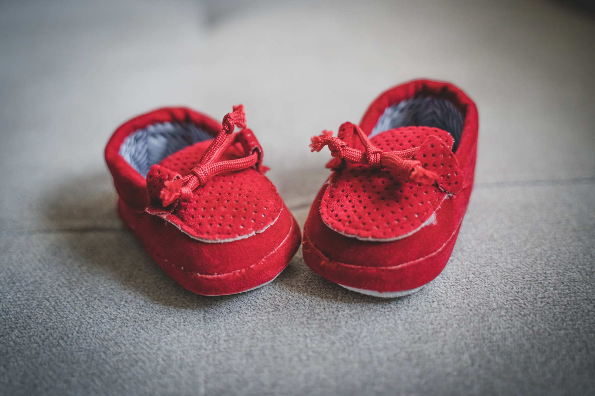 Dwa czerwone, zamszowe buciki niemowlęce z dziurkowanym wzorem i sznurowadłami, leżące na szarym tle.