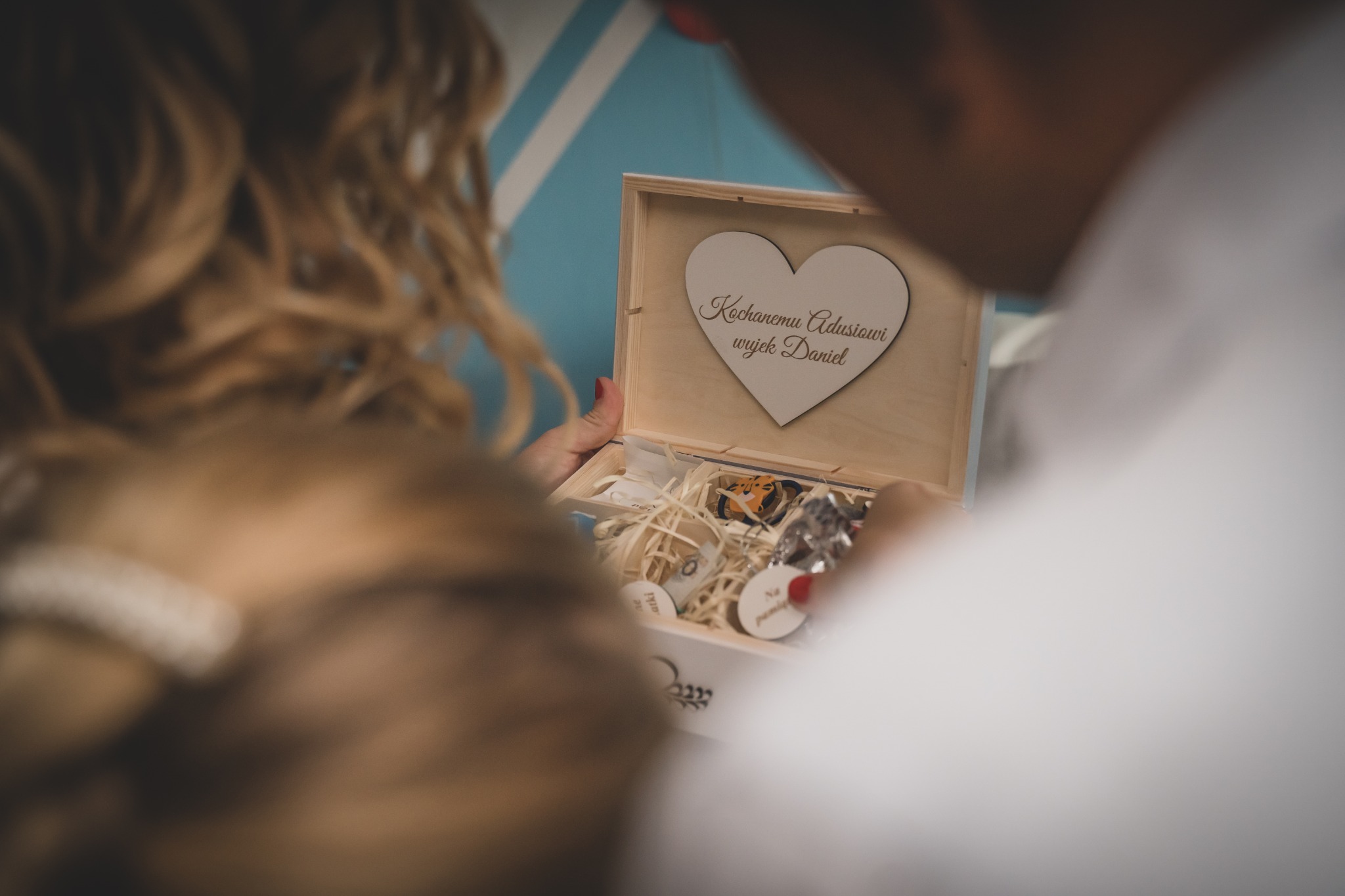 Otwarta drewniana skrzynka z wygrawerowanym sercem i napisem 'Kochanemu Adusiowi wujek Daniel', wypełniona pamiątkami i ozdobnym siankiem, trzymana przez parę osób.