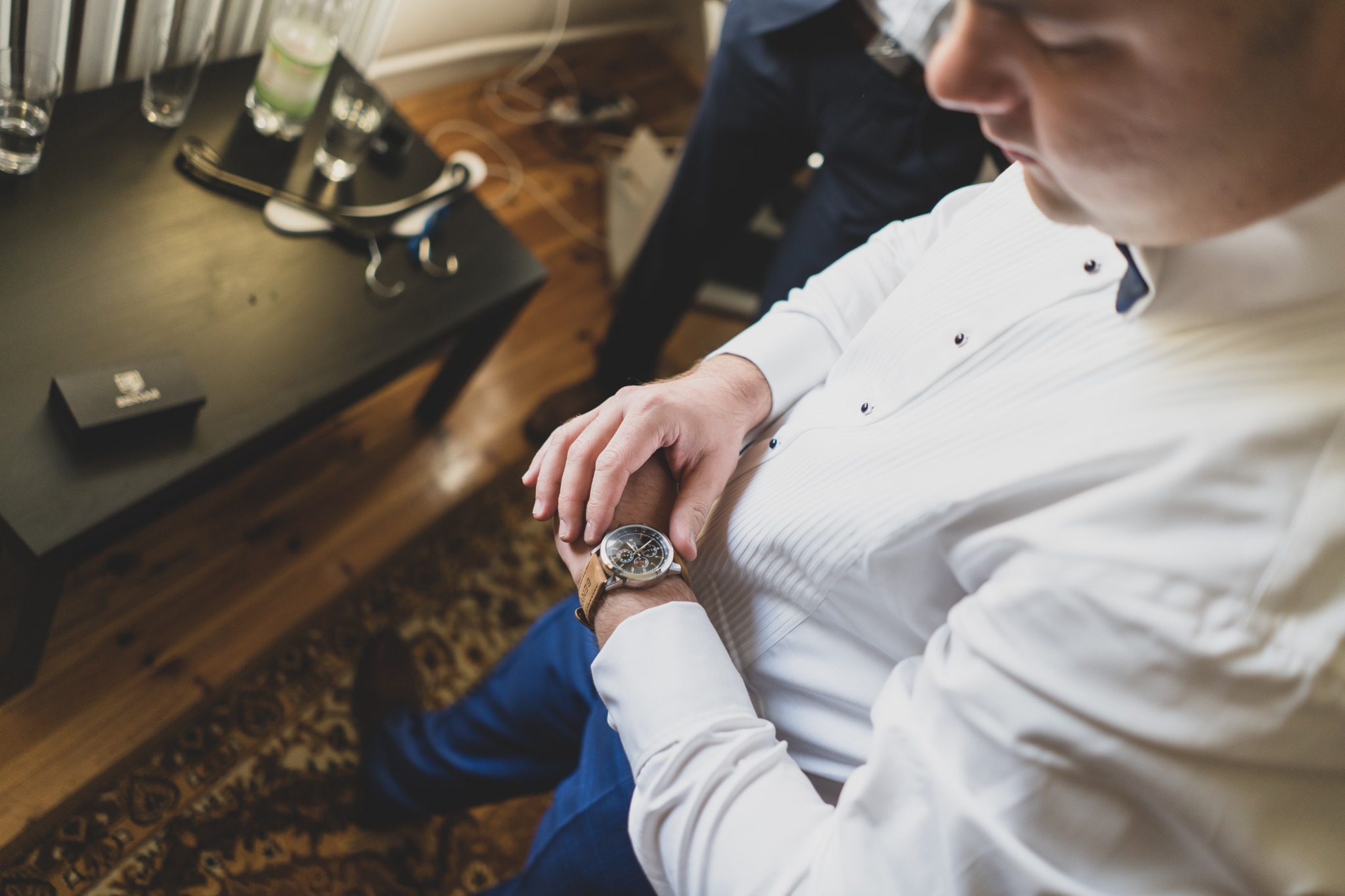 Mężczyzna w białej koszuli z mankietami, siedząc, sprawdza czas na zegarku z brązowym paskiem, widoczny fragment wnętrza z dywanem w orientalne wzory i stolikiem z wizytówką.