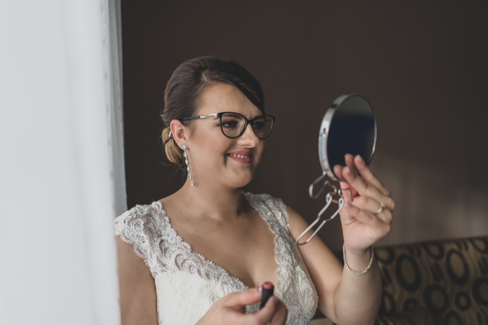 Uśmiechnięta panna młoda w okularach, poprawia makijaż ust szminką, spoglądając w okrągłe lusterko ręczne, w tle widoczne firanki i fragment sofy.