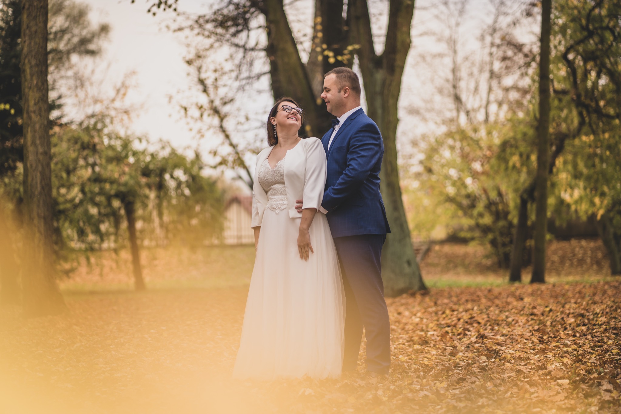 Para młoda, panna młoda w okularach i sukni ślubnej z koronkową górą, pan młody w niebieskim garniturze, stoją na tle parku jesienią, pokrytego opadłymi liśćmi.