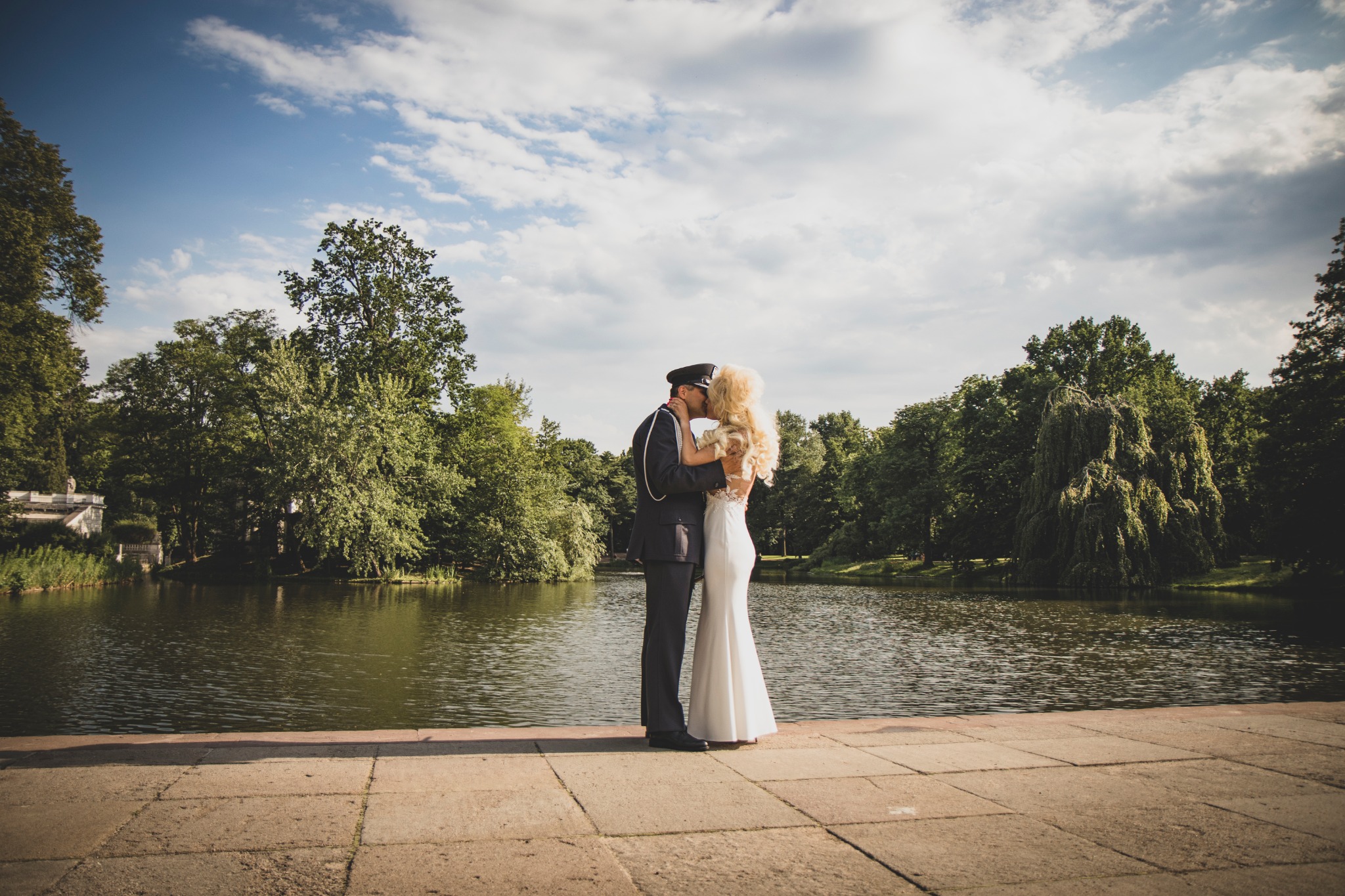 Pocałunek pary młodej w mundurze i białej sukni na tle jeziora i zieleni w parku, pod błękitnym niebem z chmurami.