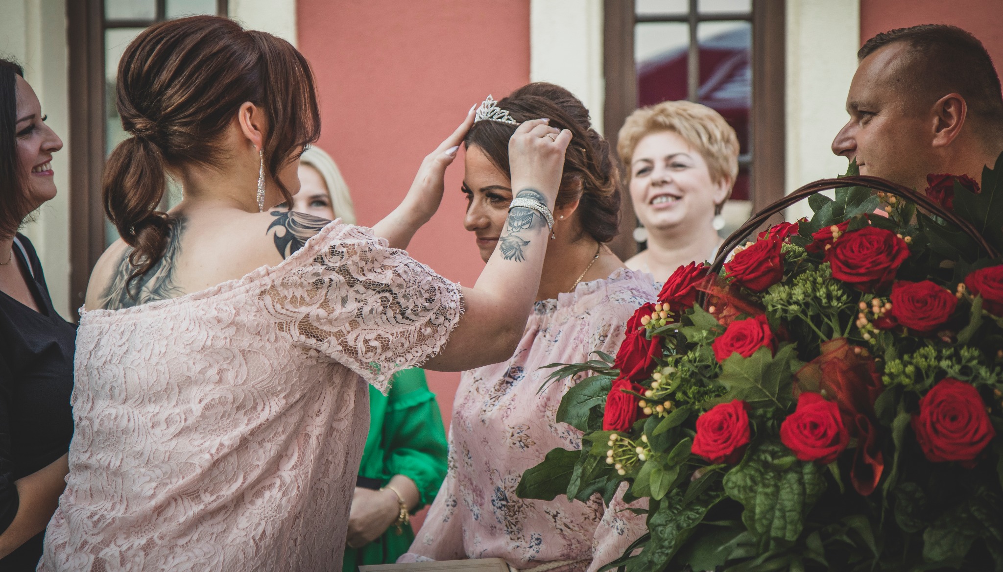 Kobieta w różowej sukience zakłada tiarę innej kobiecie, w tle kosz pełen czerwonych róż i uśmiechnięci goście.