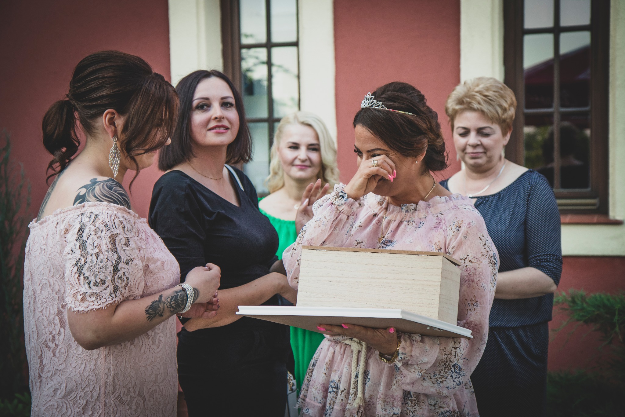 Wzruszona panna młoda w koronie, otoczona druhnami, trzyma drewniane pudełko z pamiątkami ślubnymi na tle czerwonej ściany budynku z oknami.