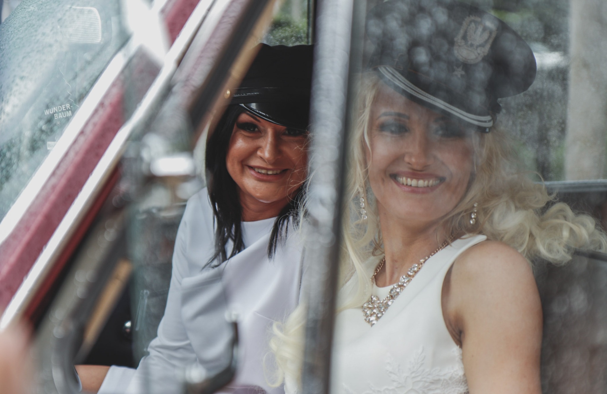 Dwie uśmiechnięte kobiety w stylizowanych strojach i czapkach siedzą w samochodzie retro, widoczne przez zaparowaną szybę z naklejką 'Wunder-Baum'.