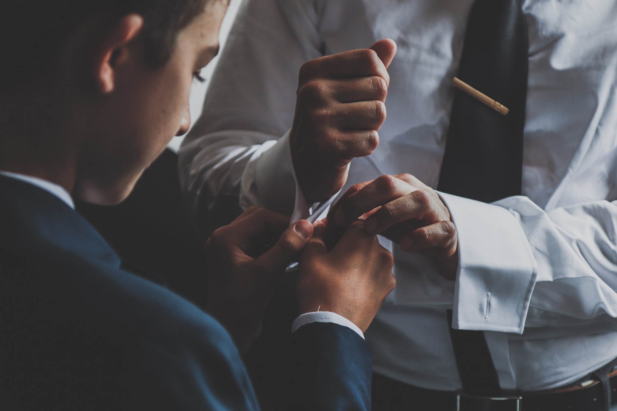 Mężczyzna w białej koszuli zapinający mankiet, widoczny fragment ciemnego garnituru, czarny krawat z ozdobną spinką.