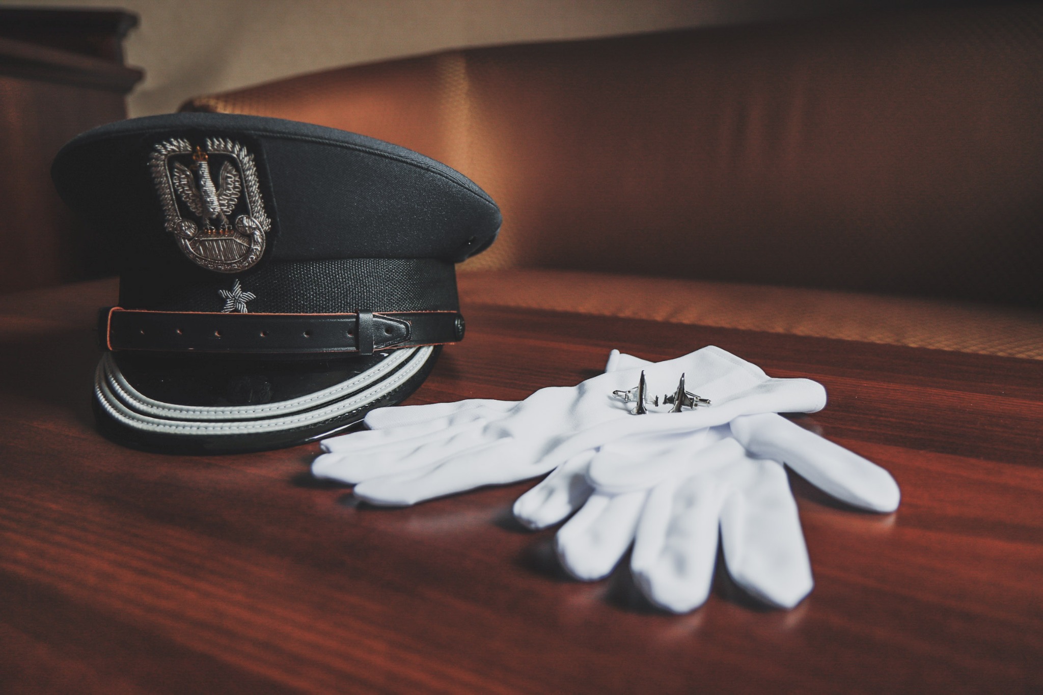 Czarny kapelusz galowy z godłem, białe rękawiczki i spinki do mankietów w kształcie samolotów na ciemnobrązowym drewnianym stole.
